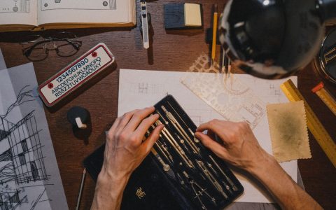 Overhead view of an architect's hands working on blueprints with drafting tools on a wooden desk.