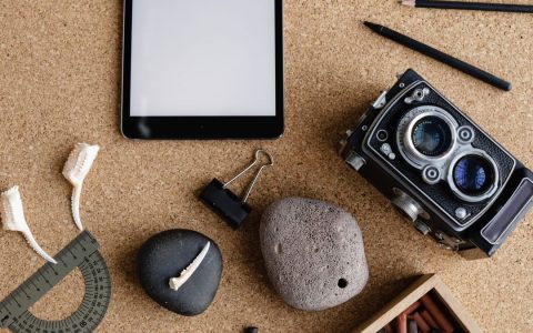 A flatlay of vintage camera, tablet, pencils, stones, and art supplies on a cork desk.