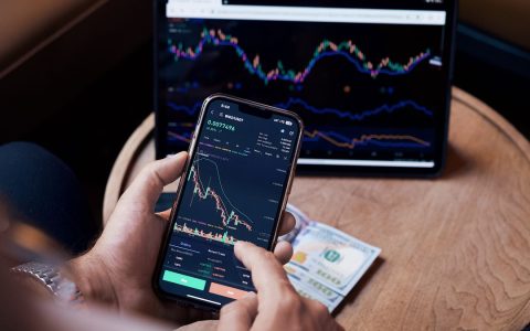 Close-up of a person trading stocks using a smartphone and a tablet.