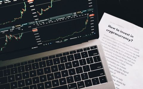 Close-up of a laptop displaying stock market graphs with a paper on investing in cryptocurrency, indoors.