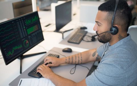 Man in a headset analyzing stock data on a computer monitor at his workspace.