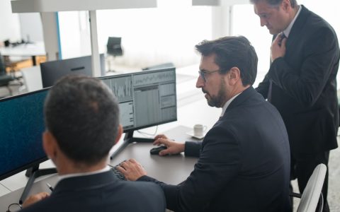 Colleagues in a modern office reviewing stock market trends and data on multiple screens.