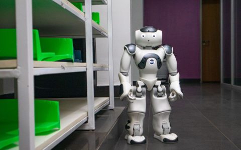 A humanoid robot standing in a modern corridor with shelves, representing futuristic technology.