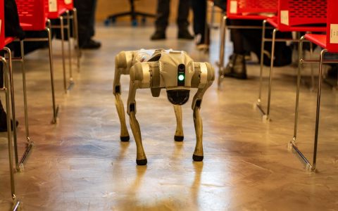 A robotic dog navigates an indoor setting amidst red chairs, showcasing technology in modern environments.