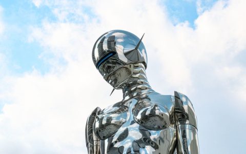 A sleek chrome robot sculpture stands against a bright blue sky background.