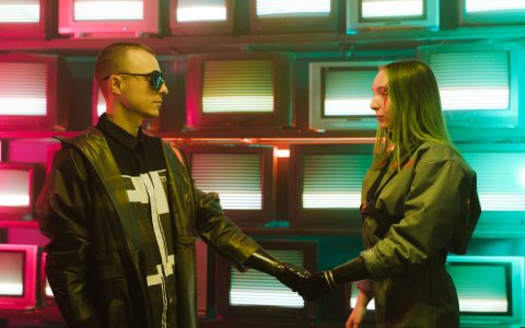 Two individuals with prosthetic arms engage in a handshake amidst a vibrant, retro television backdrop.