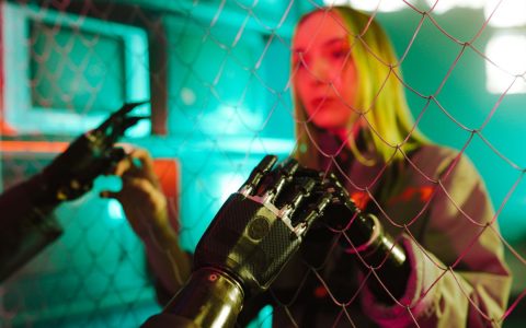 A woman interacts with a robotic hand through a fence under neon lights.