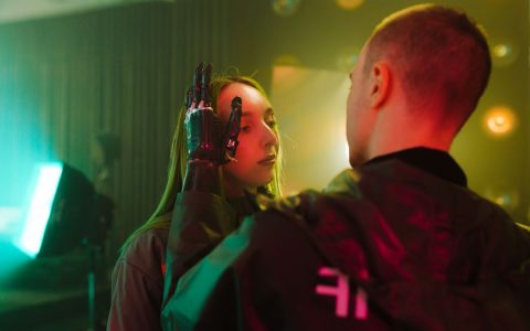 A man with a prosthetic hand gently touches a woman's face in a cyberpunk setting.