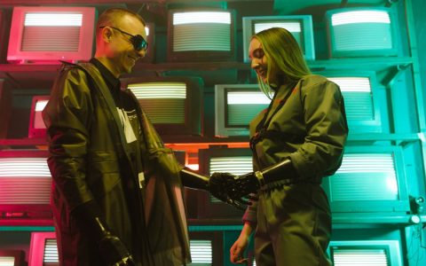 Futuristic scene of two adults shaking hands with prosthetics against a retro TV backdrop.