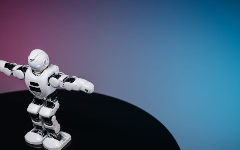 A white humanoid toy robot standing on a reflective black surface in a studio setting with a blue and pink gradient background.
