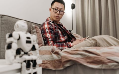 Asian man in plaid shirt sitting on bed, observing a robot toy in a cozy bedroom setting.