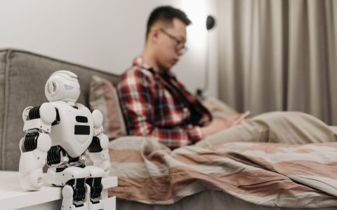 A modern bedroom featuring a robot on a bedside table and a man sitting on a bed.