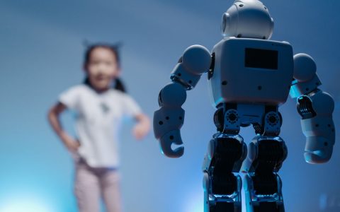 A young girl playfully interacts with a humanoid robot in a futuristic indoor environment featuring soft blue lighting.