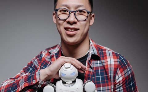 Asian man smiling, holding a small toy robot during a studio shoot. Indoors, with eyeglasses and plaid shirt.