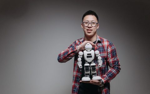 Asian man with eyeglasses holding a toy robot in a studio with a gray background.