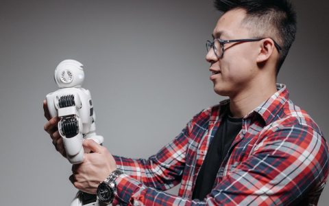 Asian man with glasses in plaid shirt holding a robot toy indoors. Studio shot.