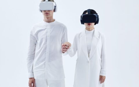 Two people in white attire wearing virtual reality headsets and holding hands, embracing technology.