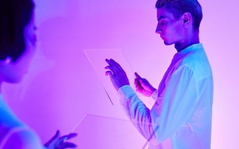 Two individuals engaging with futuristic transparent touch screens in a vibrant purple-lit studio.
