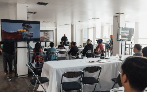 Speaker presenting to a diverse audience at a modern indoor conference setting.