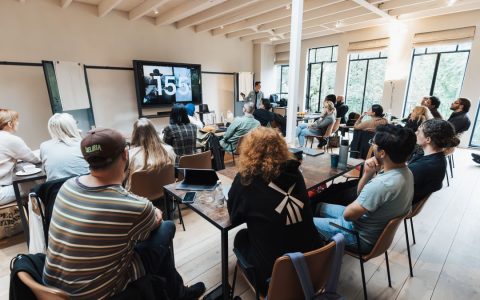 Diverse group attending a seminar in a modern wooden interior, focusing on a large screen presentation.