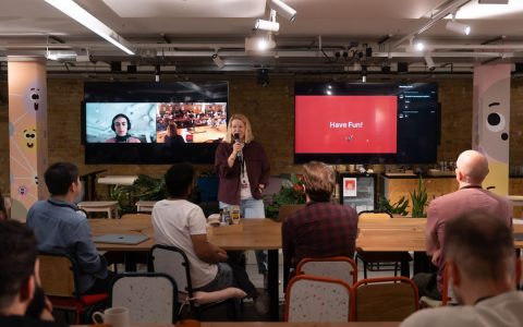 A speaker engages with an audience in a modern conference setup. Interactive screens enhance the presentation.