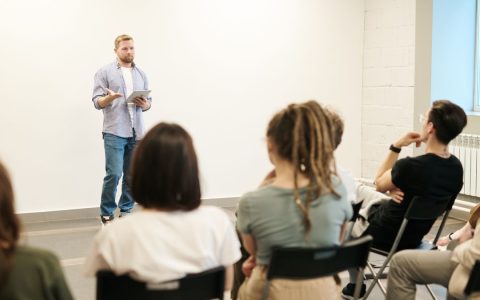 Man presenting with tablet to a small seated audience in a modern indoor setting.