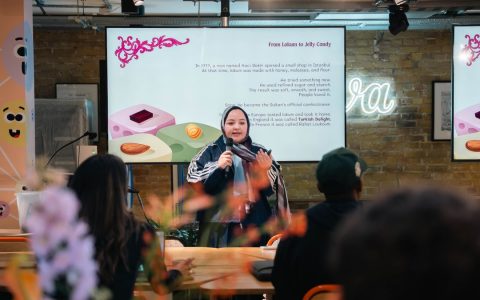 A woman presents in a modern creative workspace with a large digital screen and neon sign.