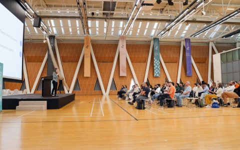 Large indoor conference with audience focused on a speaker presenting from a podium.