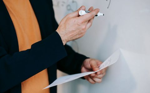 Close-up of a teacher explaining math concepts on a whiteboard while holding notes.