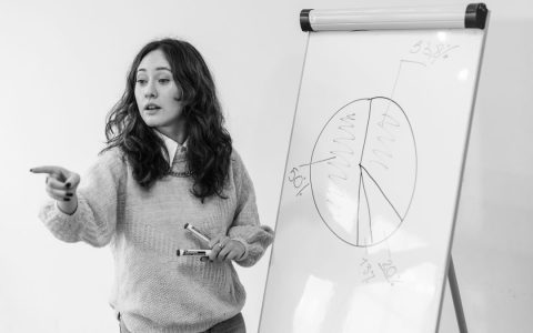 A woman in a sweater giving a presentation with a graph on a whiteboard, pointing enthusiastically.