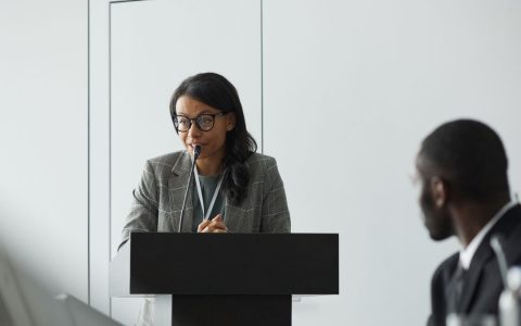 African American businesswoman delivering a speech at a conference podium with a colleague listening.