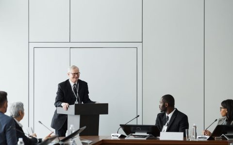 Formal business meeting with a diverse group and speaker at a podium in a conference room.