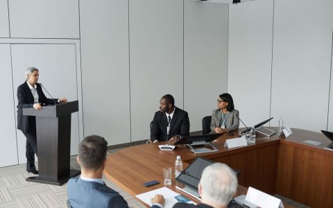 Diverse group of professionals in a formal meeting setting with a speaker at the podium.
