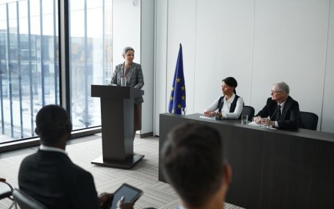 Professional conference setting with a speaker at a podium and attentive audience in a modern meeting room.