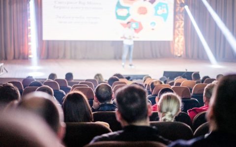 Audience attentively watching a presentation in a conference setting, with stage lights and speaker visible.